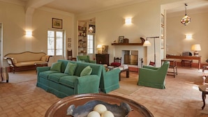 Amplia sala de estar con un sofá verde, un sillón a rayas y una mesa de centro de madera. Cuenta con una chimenea, estanterías y un comedor con mesa y sillas.