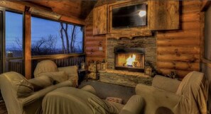 A cozy log cabin interior with a fireplace, two sofas, and a large window overlooking a forested area.