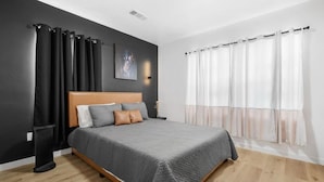 Une chambre à coucher moderne avec une tête de lit en bois, une couvre-lit grise et une grande fenêtre ornée de rideaux légers.
