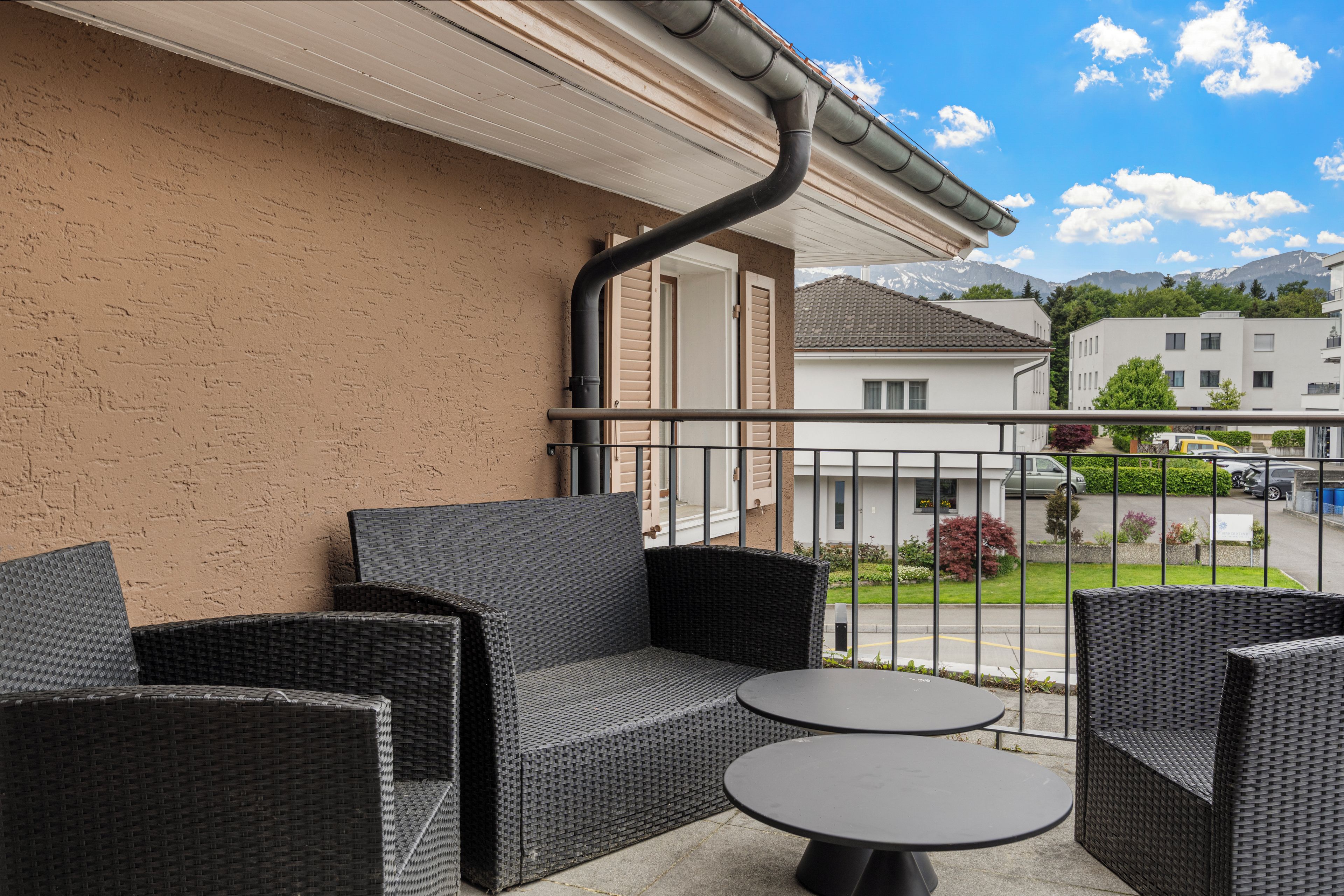 A balcony with wicker furniture and a view of a residential area with houses and mountains in the background.