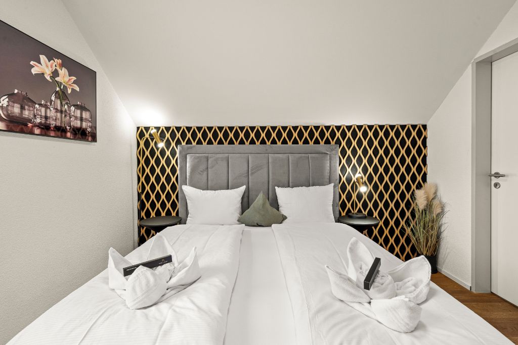 Habitación de hotel con cama, cabecero, mesita de noche y lámpara de pared.