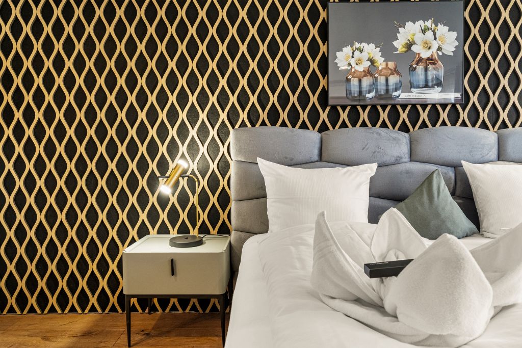 A modern bedroom with a patterned wall, a grey upholstered headboard, white bedding, and a bedside lamp.