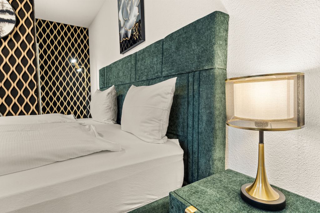 Ein ordentlich bezogenes Bett mit weißen Leinen und Kissen, ein Kopfseitenverkleidungen mit grüner Struktur und eine Tischlampe mit goldfarbener Basis.