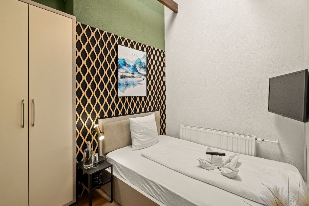 Una habitación de hotel con cama, mesita de noche, lámpara, radiador y televisor montado en la pared.