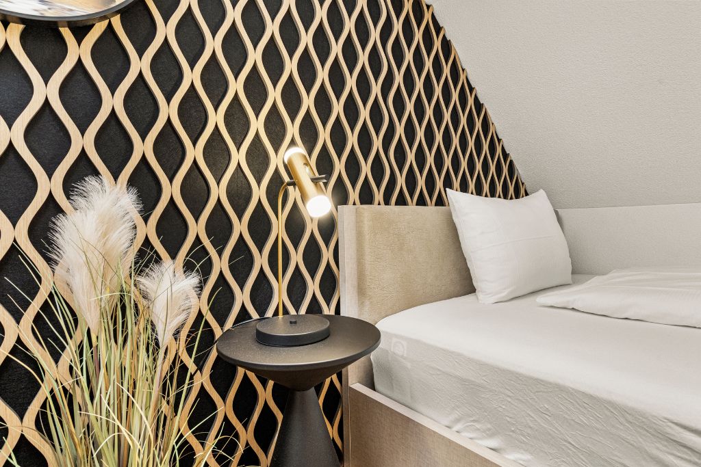 A modern bedroom with a geometric patterned wall, a bedside table with a lamp, and a bed with white bedding.