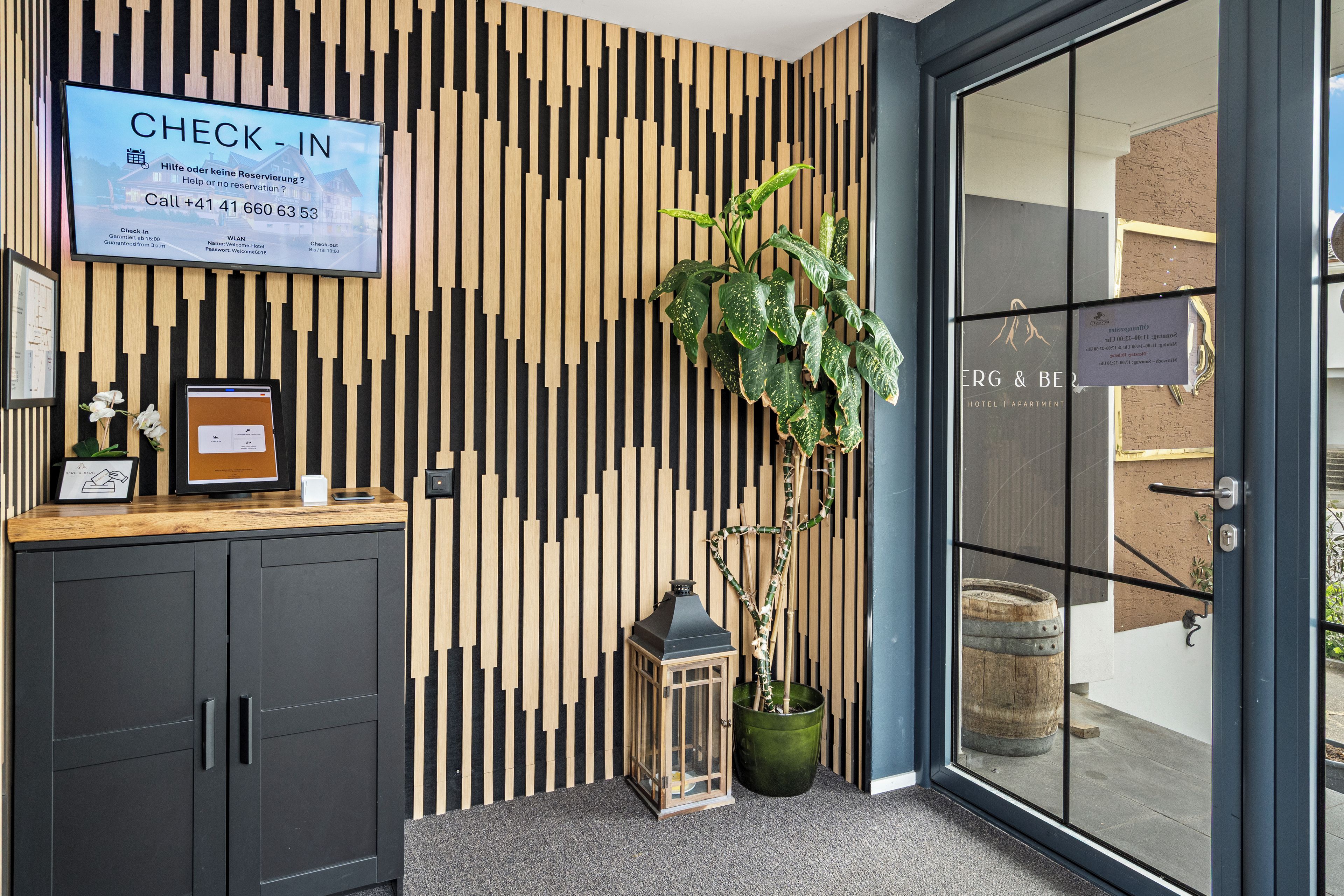 A reception desk with a check-in screen, a wooden slat wall, a potted plant, and a glass door leading to the hotel interior.