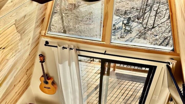 Un interior de cabaña acogedor con suelo de madera, un sofá marrón, una guitarra, un ventilador de techo y un amplio ventanal con vistas a un paisaje nevado.