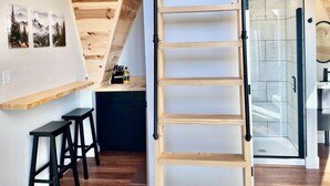 Habitación estilo loft con escalera de madera, zona de cocina y baño con ducha de vidrio.