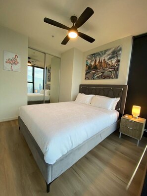 A modern bedroom with a large bed, a ceiling fan, a nightstand with a lamp, a cityscape painting, and a mirror reflecting the room.