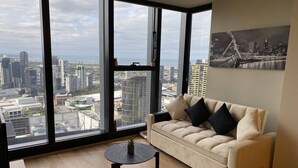 Un salon moderne avec une grande fenêtre, un canapé beige, une petite table ronde et une vue sur le paysage urbain.
