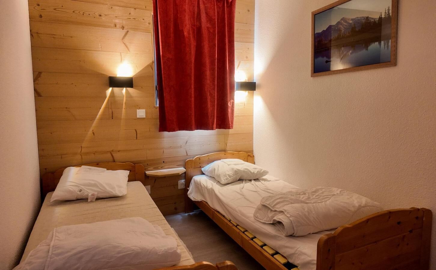 Een kleine, knusse kamer met twee houten bedden, een nachtkastje en een rood gordijn.