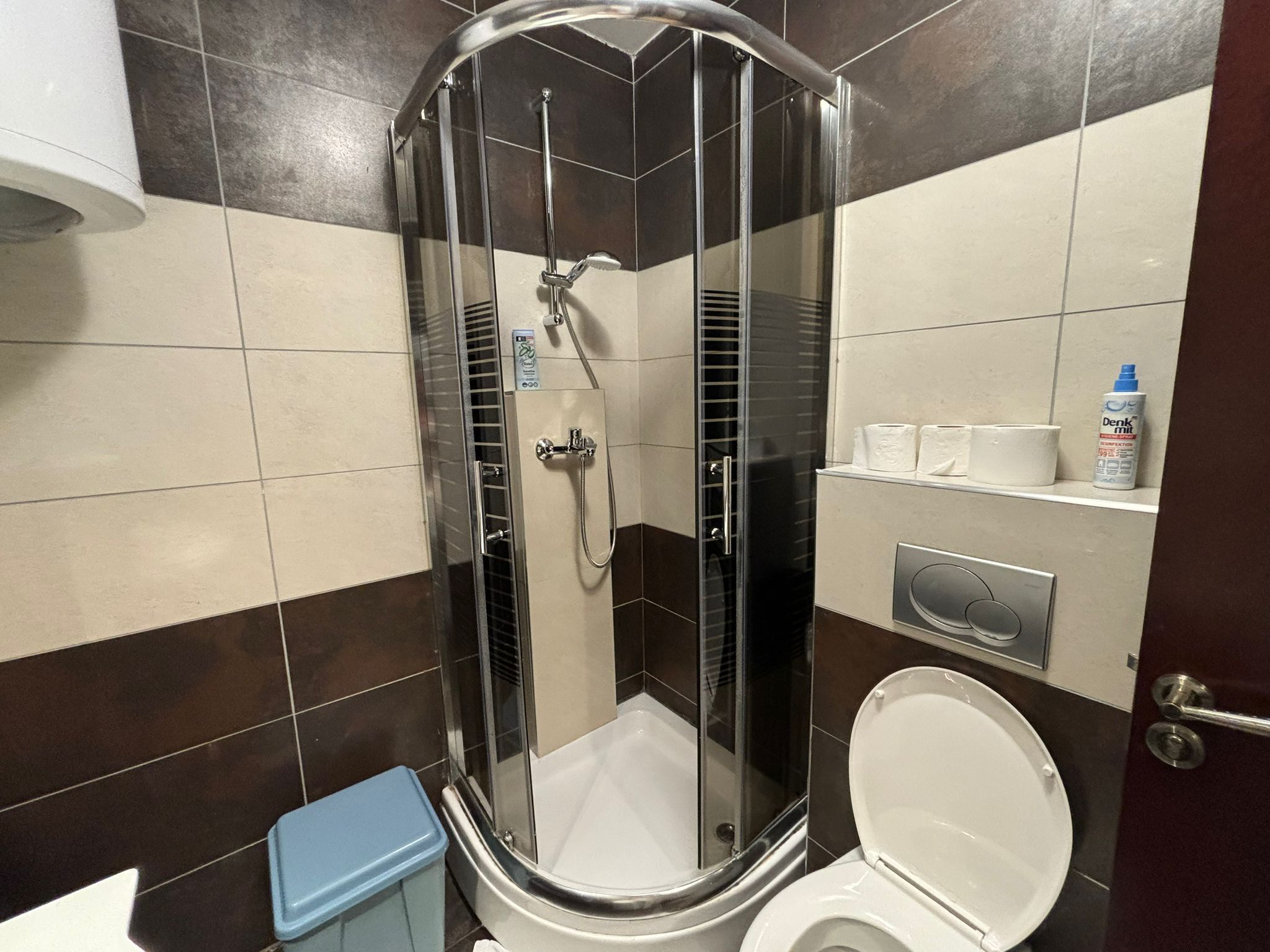 A shower enclosure with a glass door, a toilet, and a trash bin.