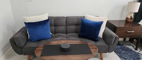 A modern living room with a grey sectional sofa, two blue and white cushions, a wooden coffee table with a black coaster, and a small wooden side table with a lamp.