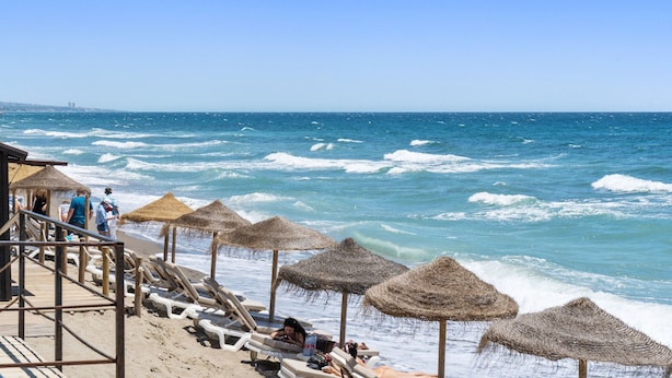 Une plage avec des parasols en chaume et des chaises longues, une terrasse en bois et l’océan en toile de fond.