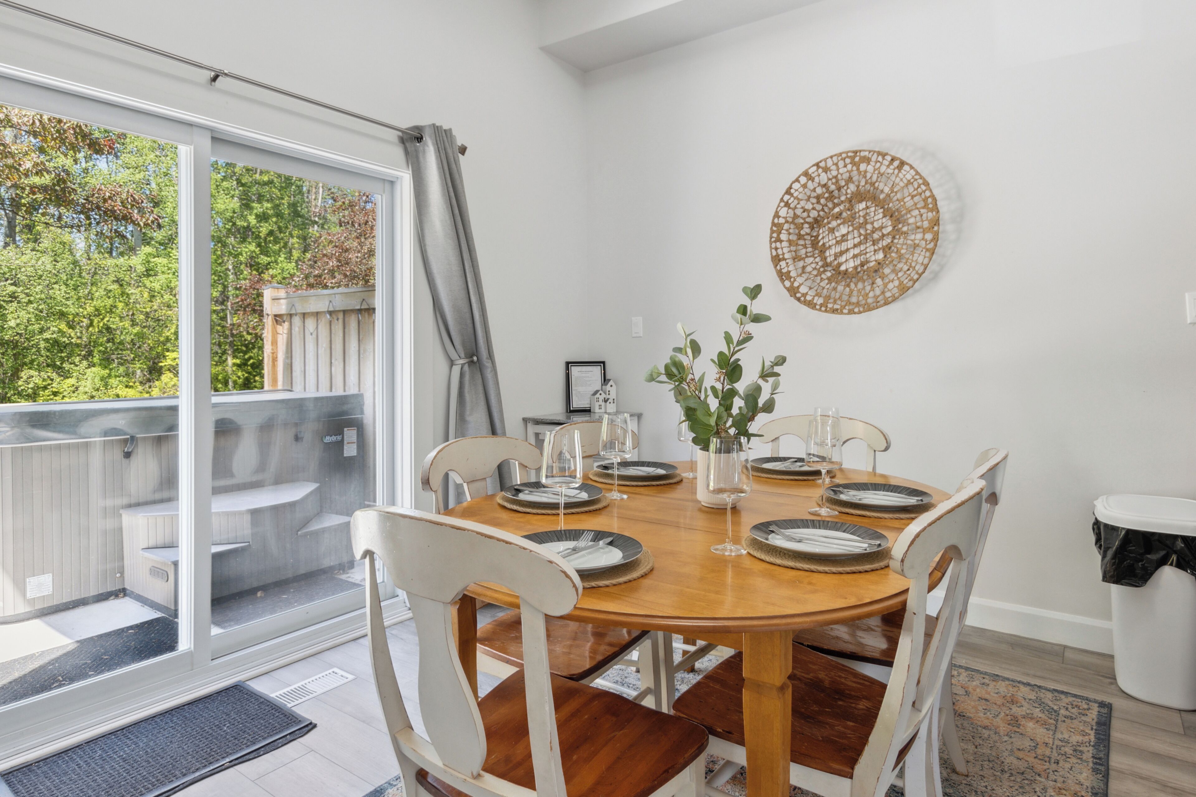 Een eetruimte met een houten tafel voor vier personen, omringd door witte stoelen. Een groot raam met uitzicht op groen is zichtbaar, en een decoratief kunstwerk hangt boven de tafel.