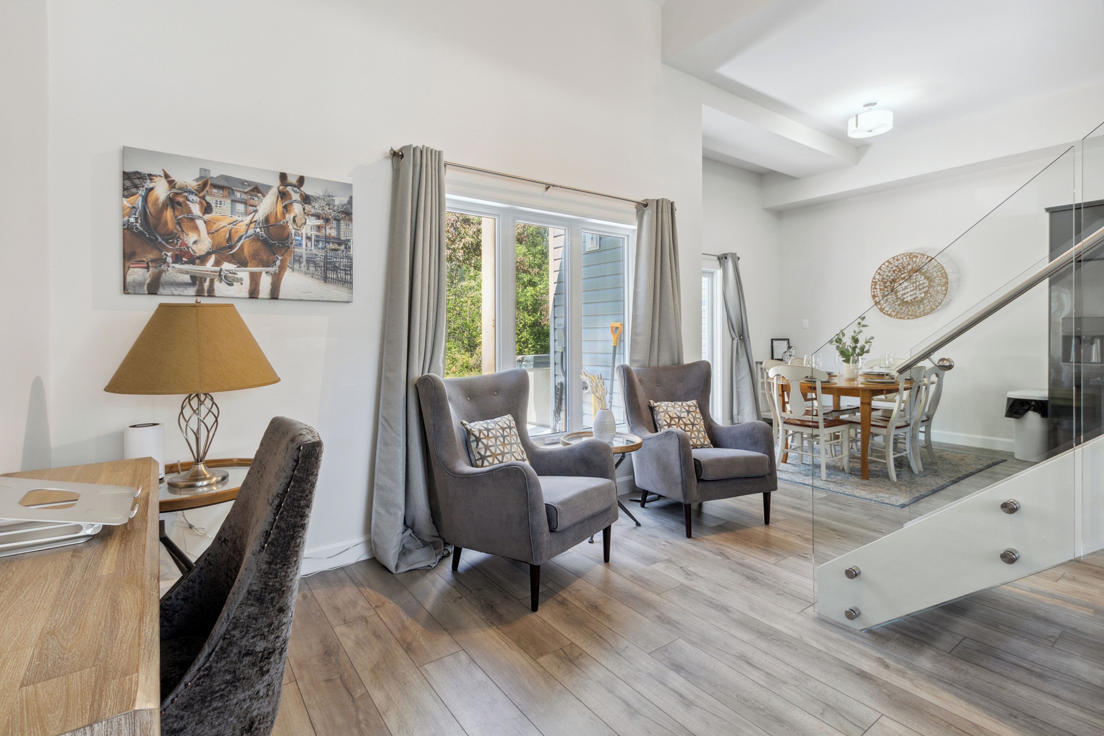 A modern living room with a wooden floor, two grey armchairs, a dining table, and a staircase with glass railings.