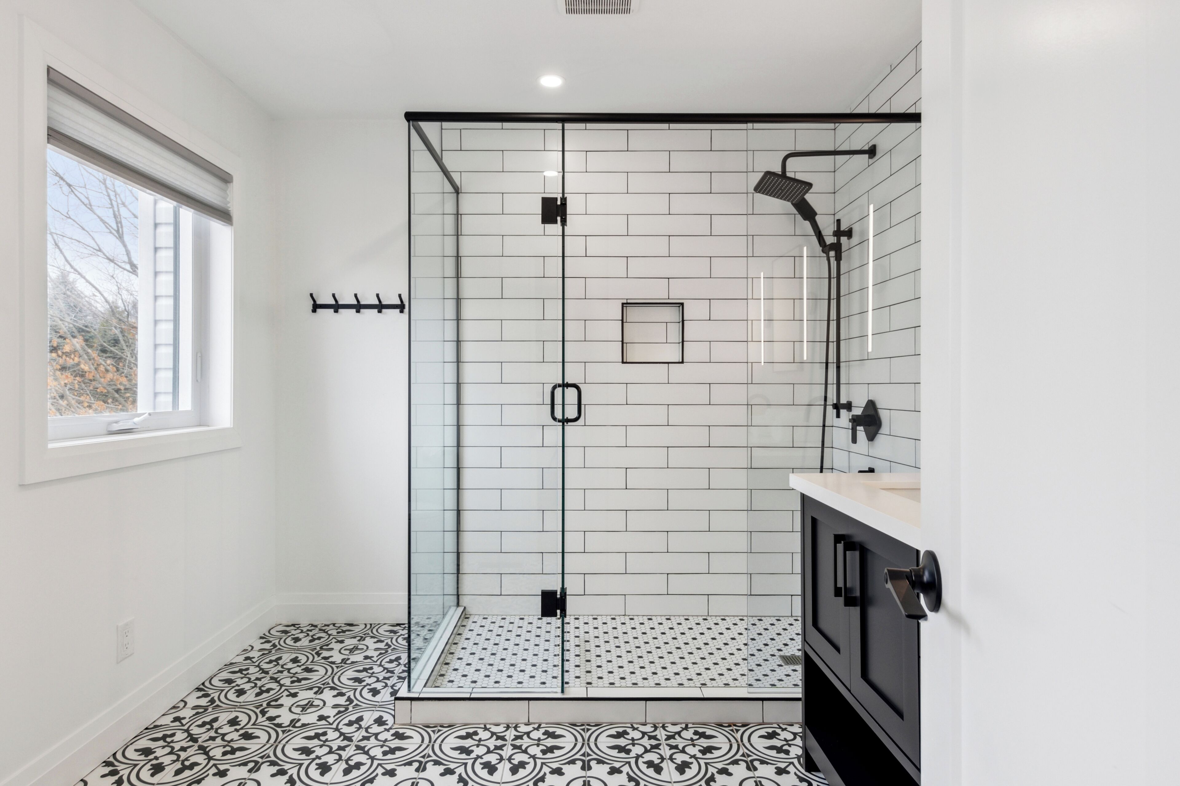 A modern bathroom with a glass-enclosed shower, white tiled walls, a patterned floor, and a window providing natural light.
