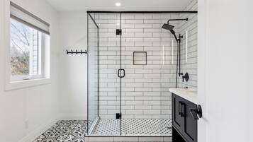 A modern bathroom with a glass-enclosed shower, white tiled walls, a patterned floor, and a window providing natural light.