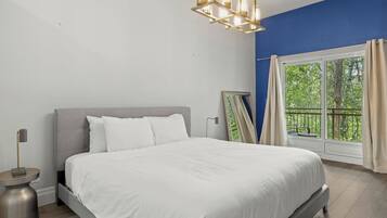 A bedroom with a bed, a nightstand, a modern chandelier, a window with curtains, and a view of greenery outside.