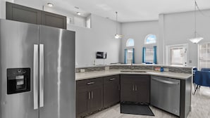 A modern kitchen with a stainless steel refrigerator, dark cabinets, a dishwasher, and a built-in oven. There is a window with blue curtains and a ceiling fan.