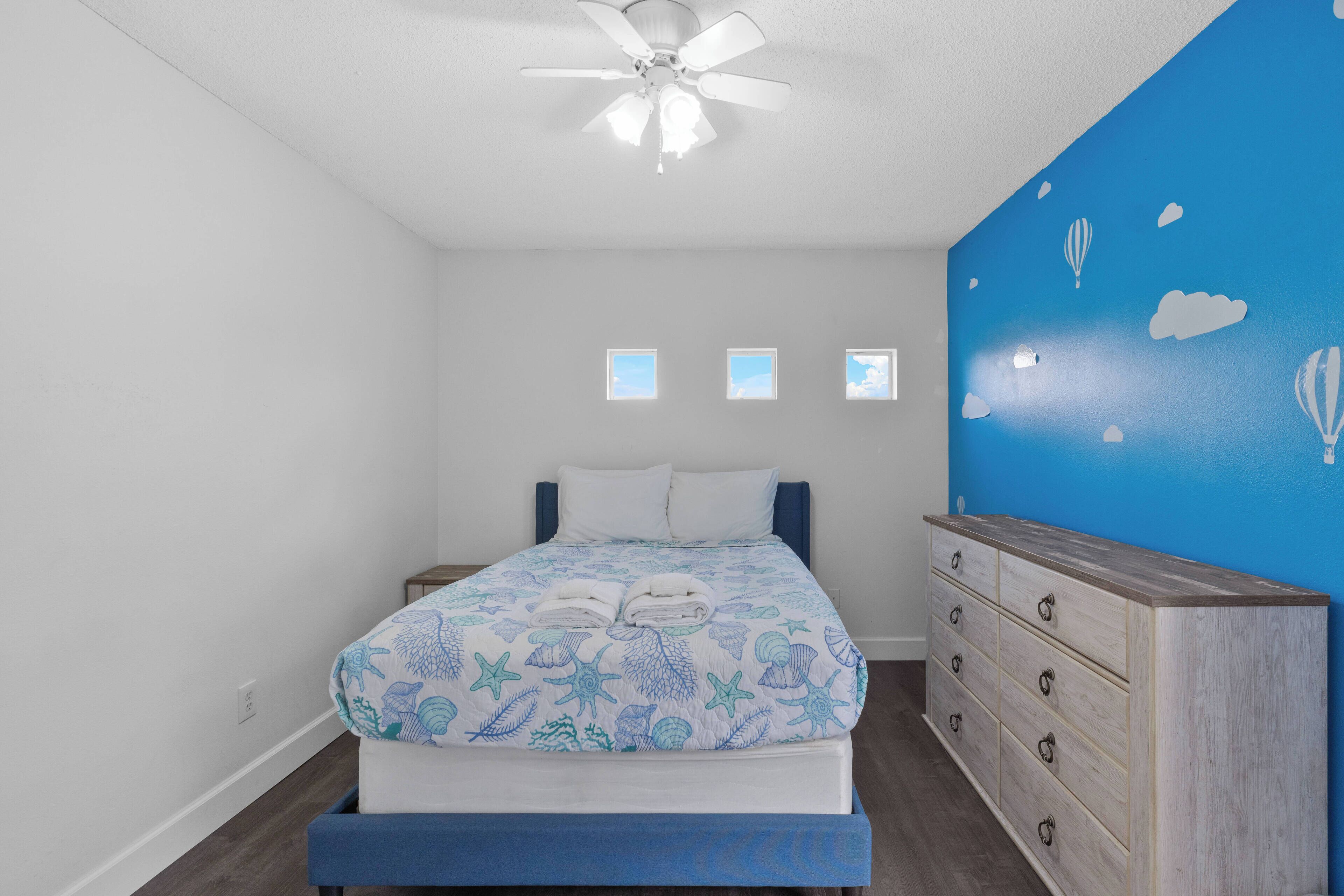 A bedroom with a bed, a dresser, a ceiling fan, and a wall with a blue mural.