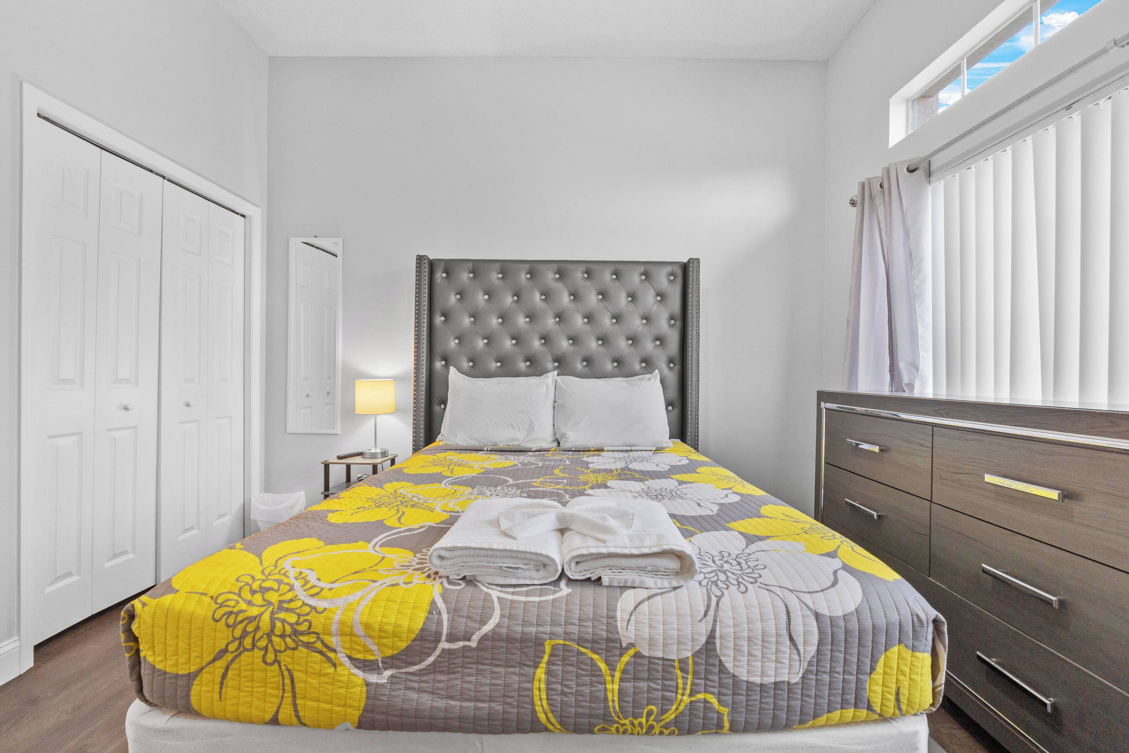 A neatly made bed with a floral bedspread, a grey tufted headboard, and two pillows. There is a wooden dresser with drawers to the right and a white door on the left.