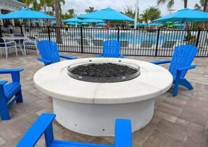 Un área de estar al aire libre circular con un brasero, rodeada de sillas y mesas azules, con una piscina y sombrillas al fondo.