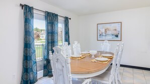 Comedor con una mesa redonda de madera, sillas blancas y vistas a una casa a través de una ventana con cortinas azules.