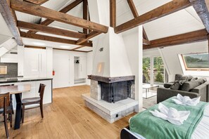 A modern loft with exposed wooden beams, a fireplace, a dining area, and a view of the outdoors.