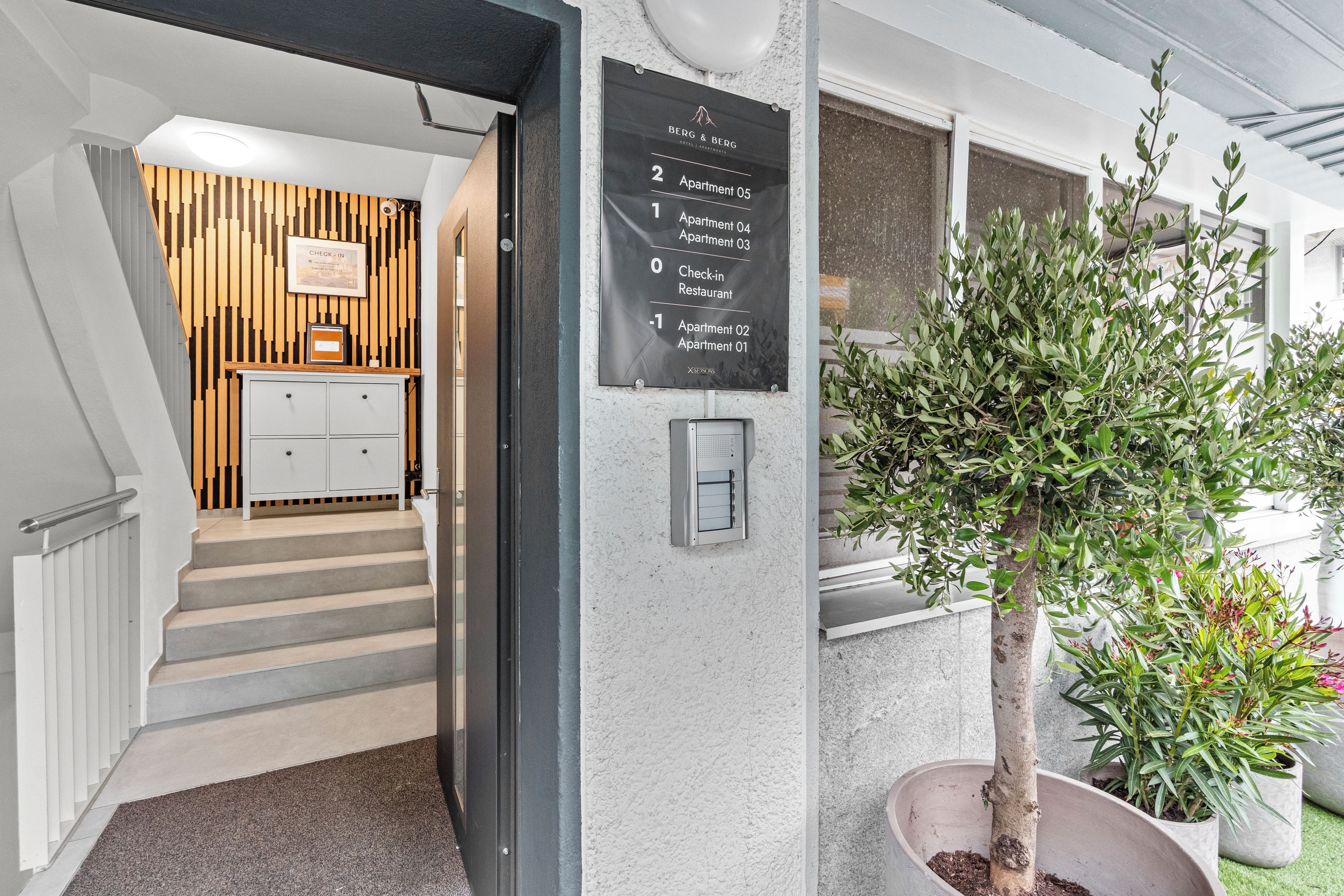 A hotel entrance with a staircase leading to a reception area, a wall-mounted directory, and a potted tree.