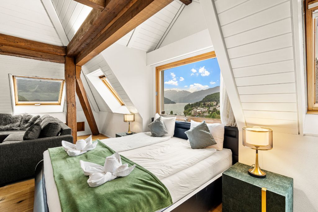 A bedroom with a bed, a nightstand, a lamp, a window with a view of mountains, and a sofa in the adjacent room.