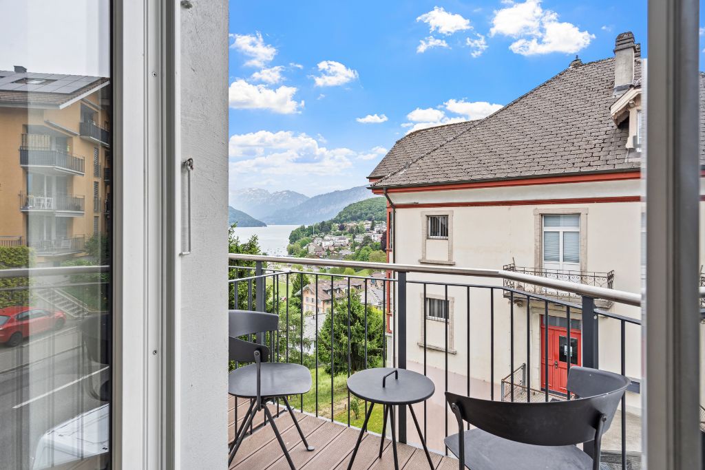 Ein Balkon mit Blick auf einen See und Berge, mit einer roten Tür, einem kleinen Tisch und einem Stuhl.