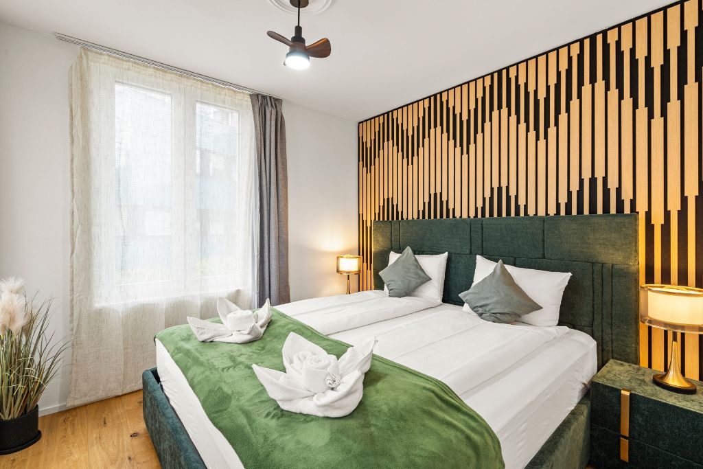 A hotel room with a bed, a headboard, a bedside table, a lamp, and a window with curtains.