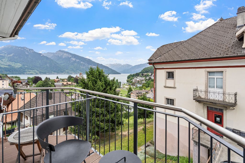 A balcony with a view of a lake and mountains, a red door, and a chair.