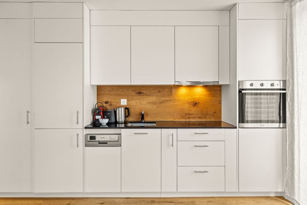 Una cocina moderna con armarios blancos, protector contra salpicaduras de madera, horno empotrado y cafetera.