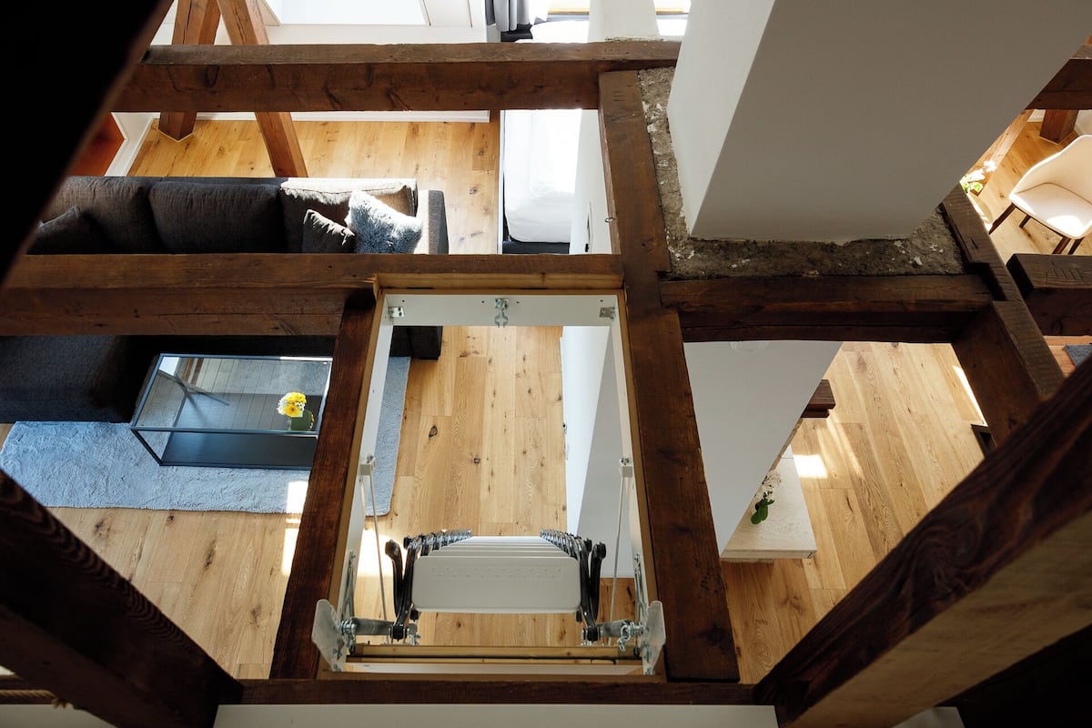 A high-ceilinged room with exposed wooden beams, a central white radiator, and a view of a living area with a sofa and a coffee table.