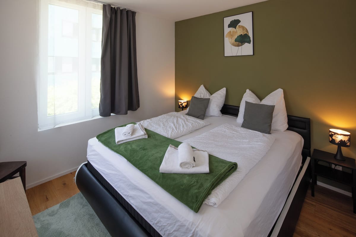 A double bed with white linens, a green throw blanket, and two towels folded on it. There is a nightstand with a lamp, a window with curtains, and a wall-mounted picture.