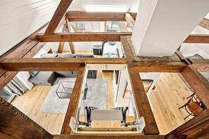 A loft-style room with exposed wooden beams, a central wooden platform, and a view of the adjacent living area.