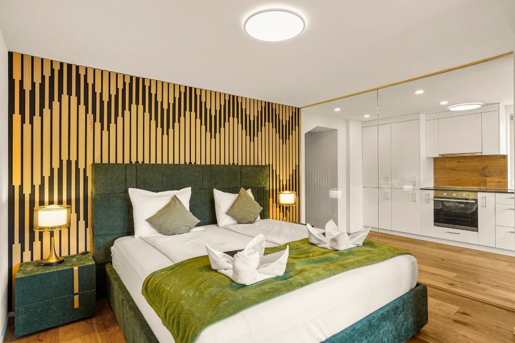 A modern hotel room with a large bed, a wooden accent wall, a kitchenette, and a separate bathroom.