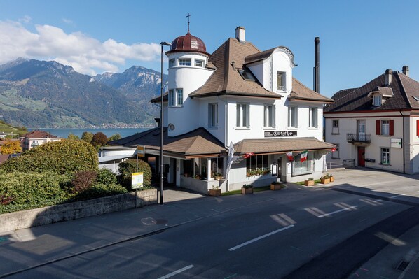 A white building with a tower, a restaurant sign, and a view of mountains. - BERG&BERG APARTMENTS-Spiez Self Check-In (Spiez)