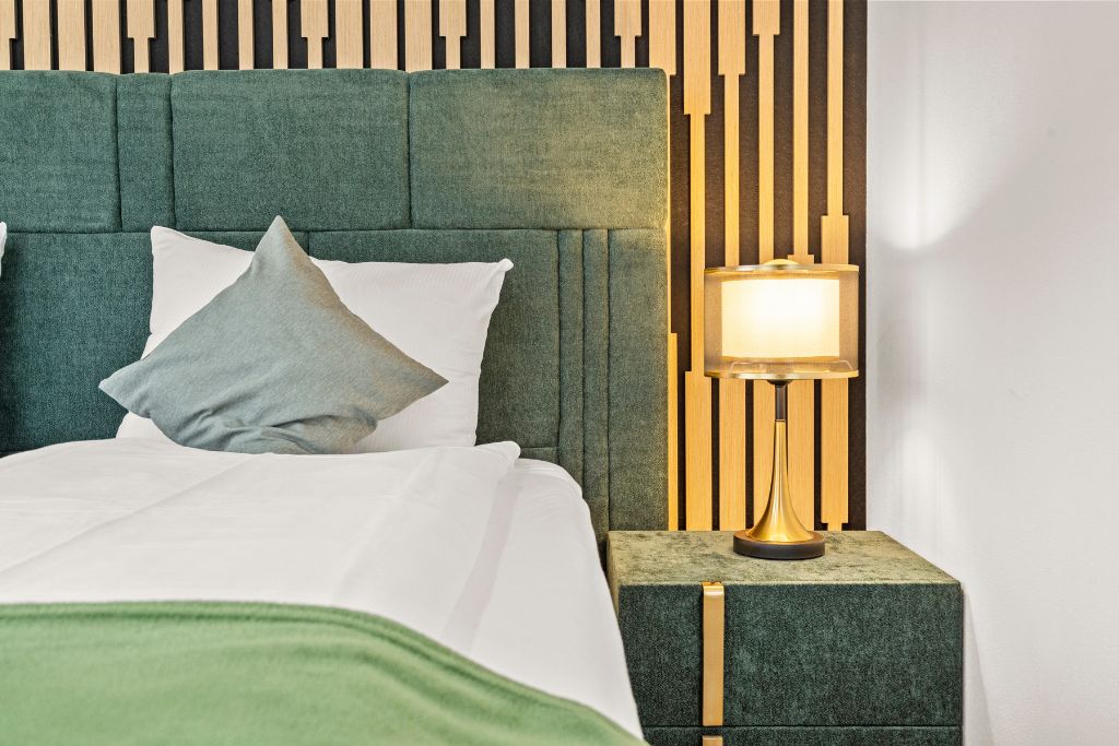 A bed with a green upholstered headboard, a bedside table with a lamp, and a wall with vertical wooden slats.