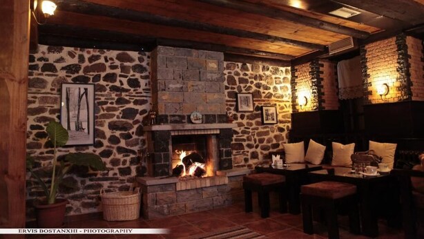 A cozy room with a stone fireplace, wooden beams, and a seating area with cushions. - Guesthouse Bujtina Leon  (Kor çë)