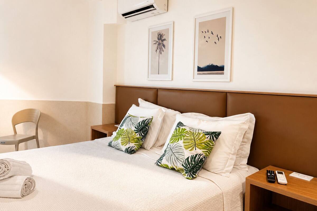 Een hotelkamer met een bed, twee sierkussens met bladmotief, een nachtkastje met telefoon en lijstjes met afbeeldingen aan de muur.