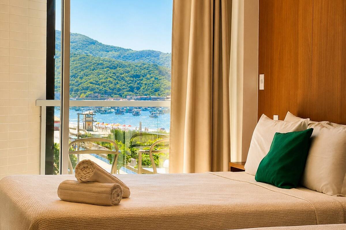 Een hotelkamer met een bed, uitzicht op een strand en bergen, en een raam met gordijnen.