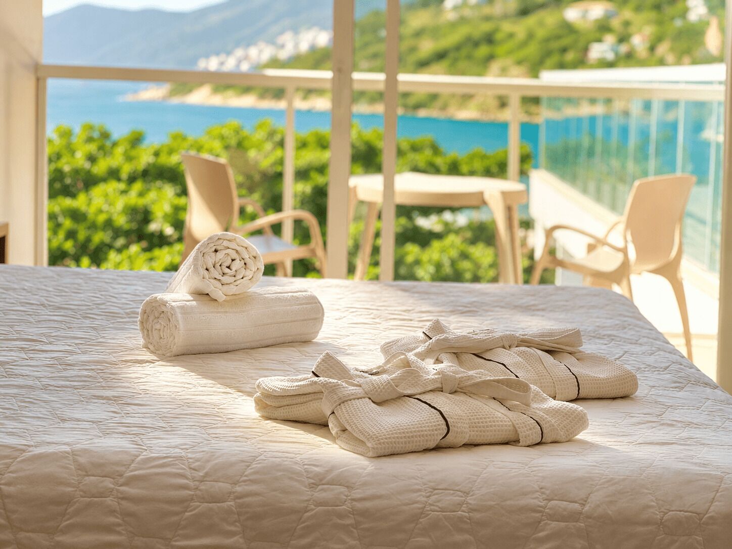 A bed with a quilted mattress, a rolled towel, and a folded blanket on a balcony with a view of greenery and a body of water.