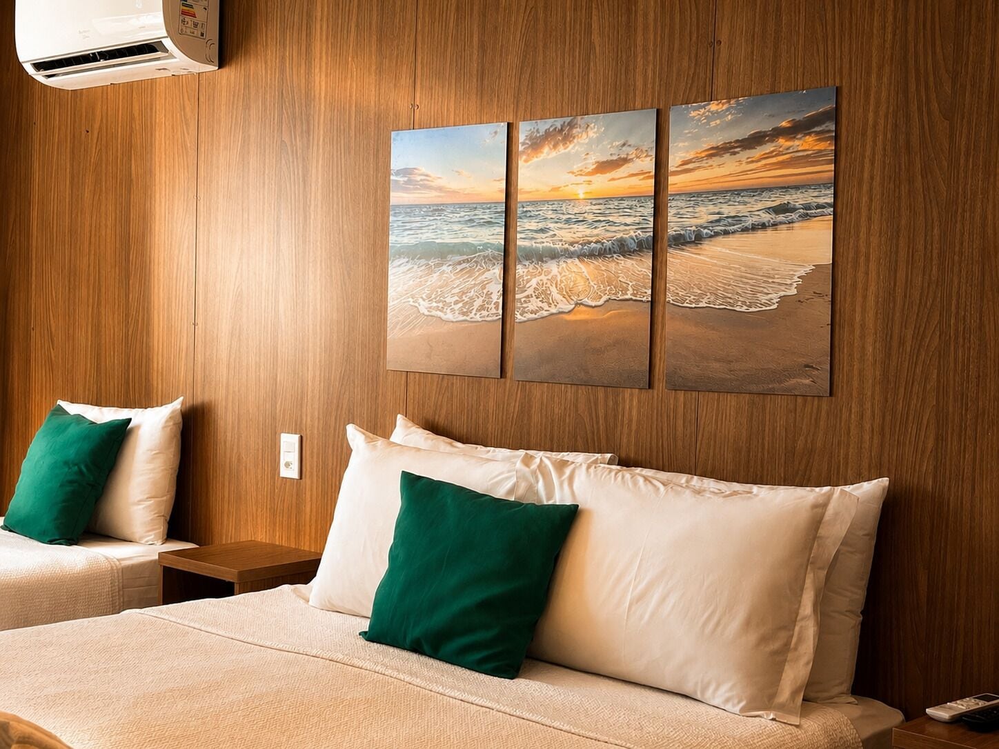Habitación de hotel con cama, dos almohadas, mesita de noche, aire acondicionado de pared y una pintura de playa al atardecer.