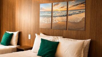 Habitación de hotel con cama, dos almohadas, mesita de noche, aire acondicionado de pared y una pintura de playa al atardecer.