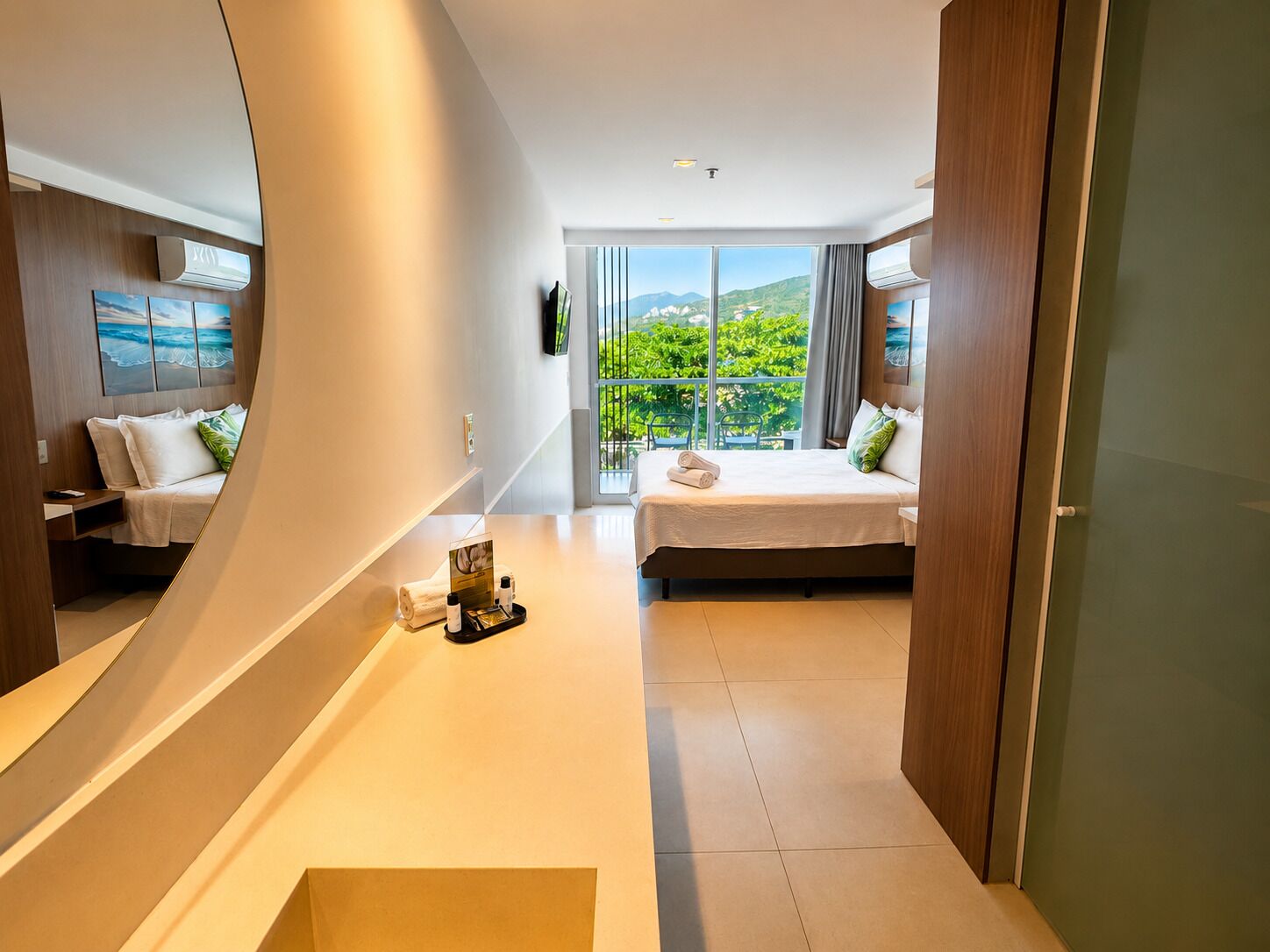 A modern hotel room with a bed, a small seating area, a large window with a view, and a mirror.