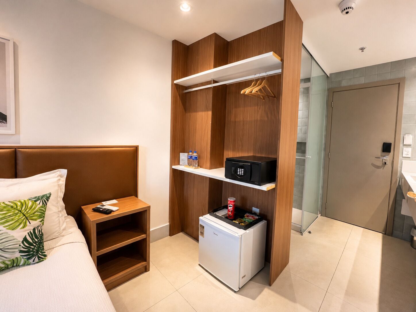 A hotel room with a bed, a small refrigerator, a minibar, a wooden wardrobe, and a bathroom visible through a glass door.