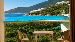 Un balcón con dos sillas y una mesa, con vistas a la costa, edificios blancos y colinas verdes.